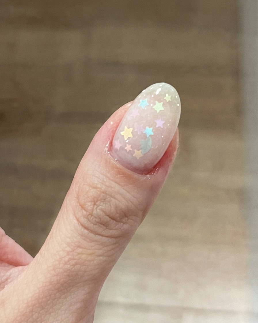 Primer plano de la uña del pulgar con una base rosa lechosa y glitters de estrellas en colores pastel colocados encima.