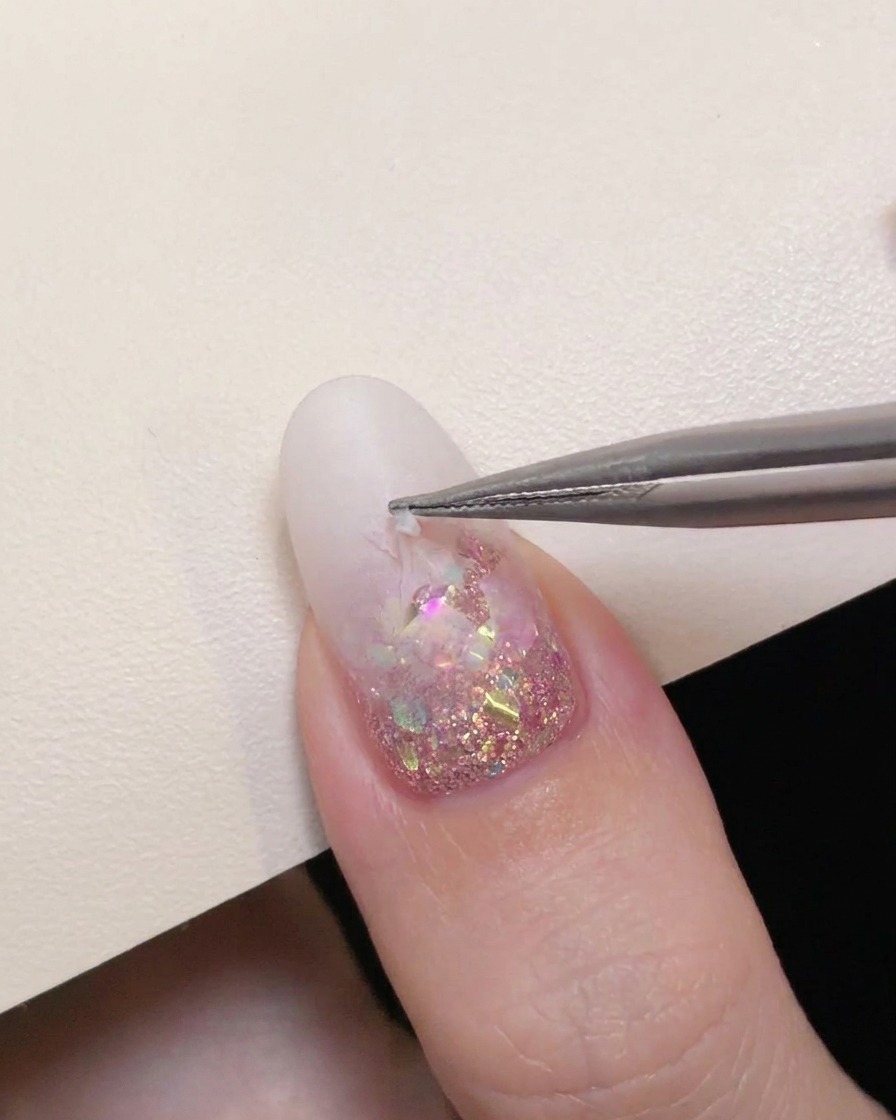 Unas pinzas de precisión retirando con cuidado una pegatina de estrella hueca de una uña con degradado blanco lechoso para revelar la purpurina rosa.