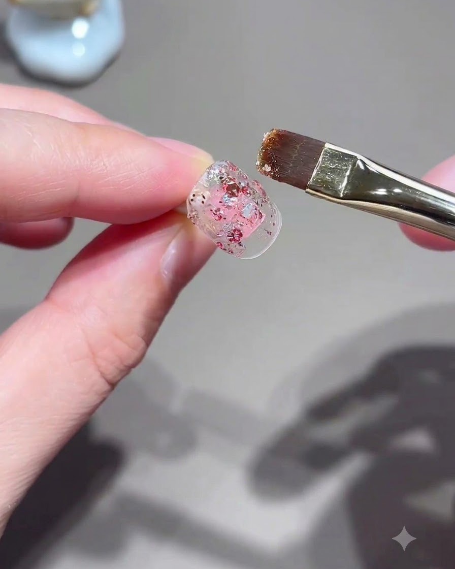 Un pincel plano aplicando gel con purpurina rosa sobre una uña.