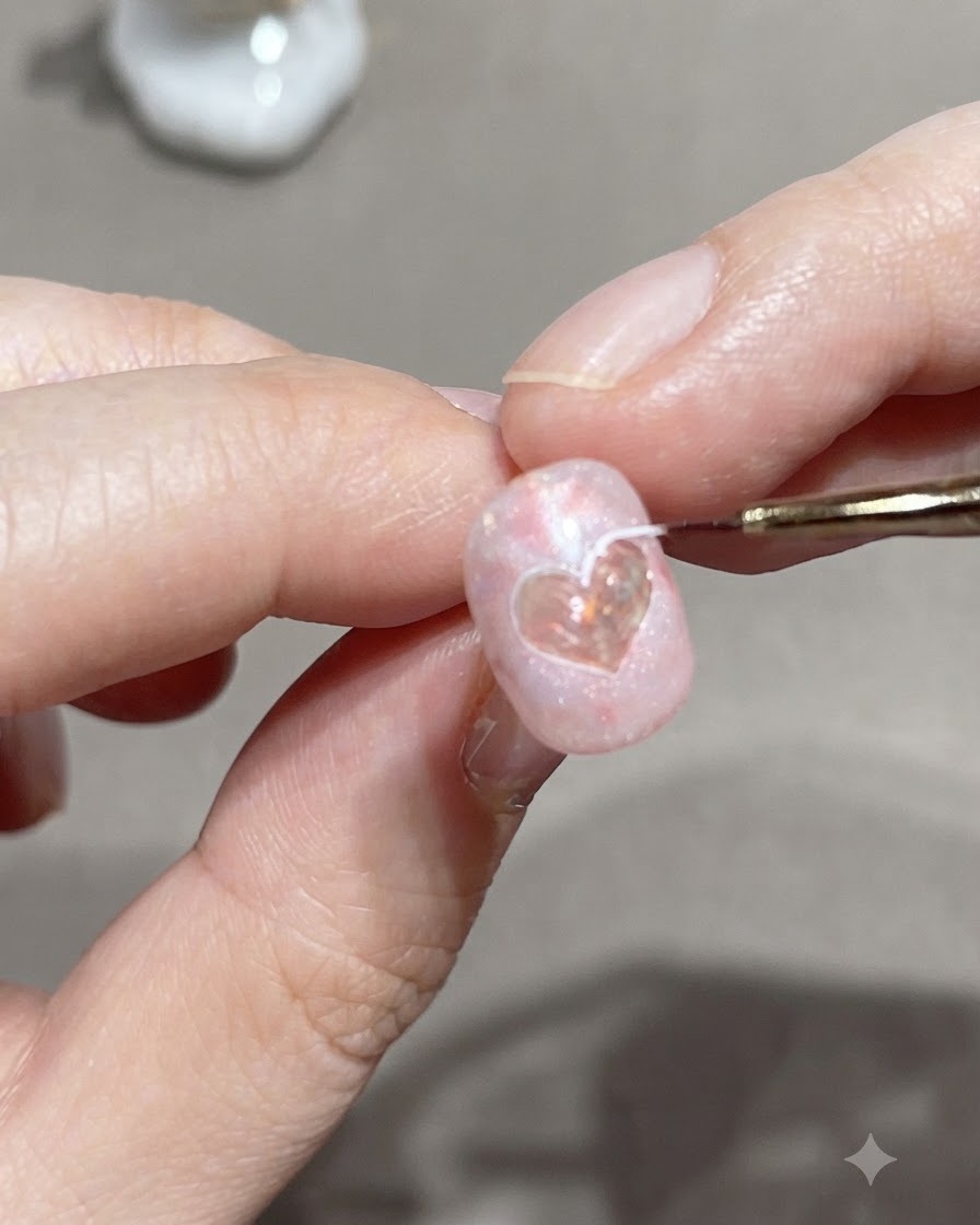 Un pincel delineador fino trazando el borde exterior de un sticker de corazón en una uña de gel rosa.