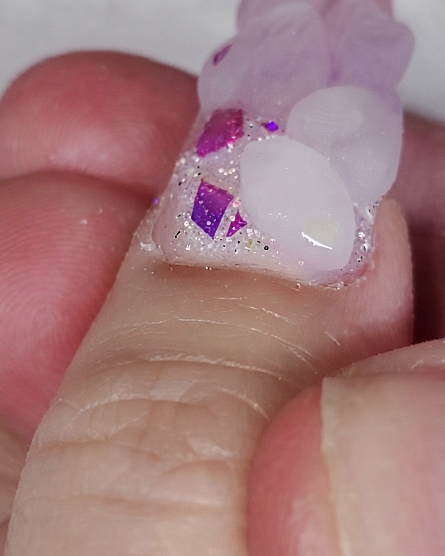 Un pincel de uñas colocando una perla acrílica rosa sobre una base con glitter para formar un corazón.