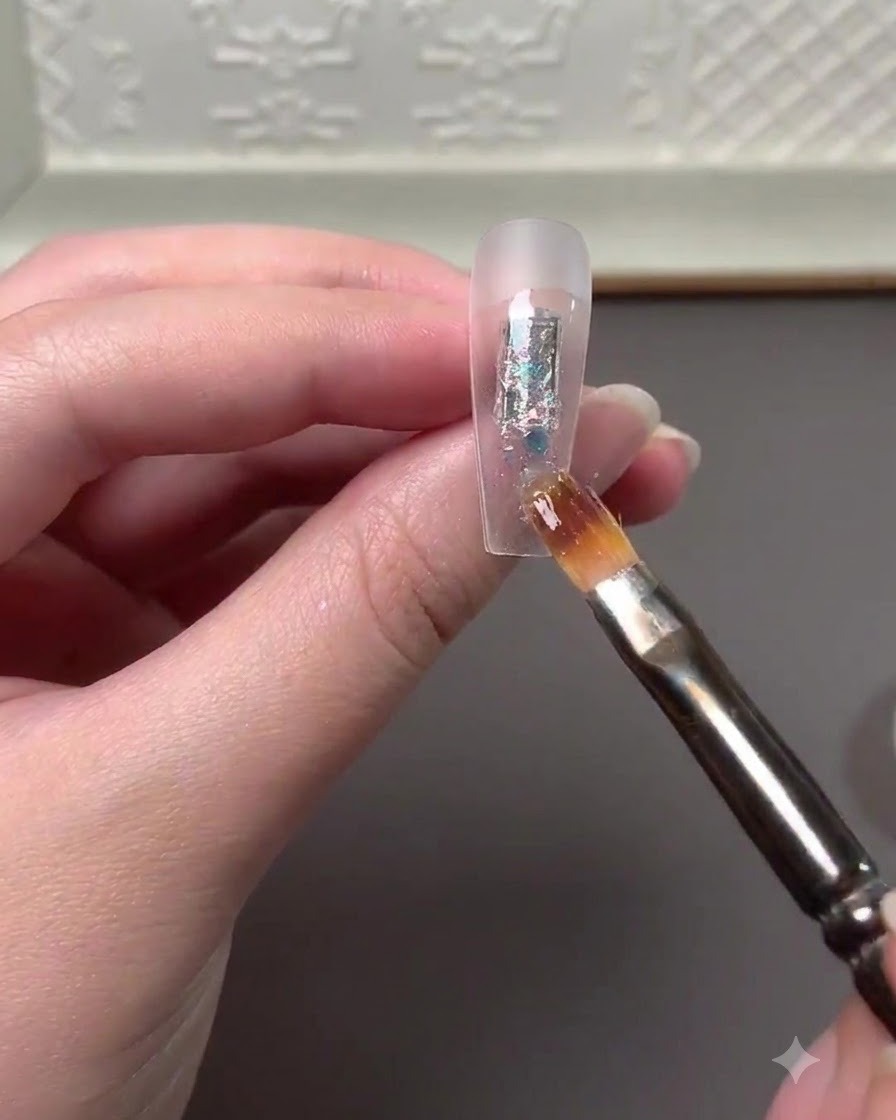 Un pincel de nail art aplicando hojuelas de glitter multicolor y polvo de alto brillo en el centro de una extensión de uña transparente.