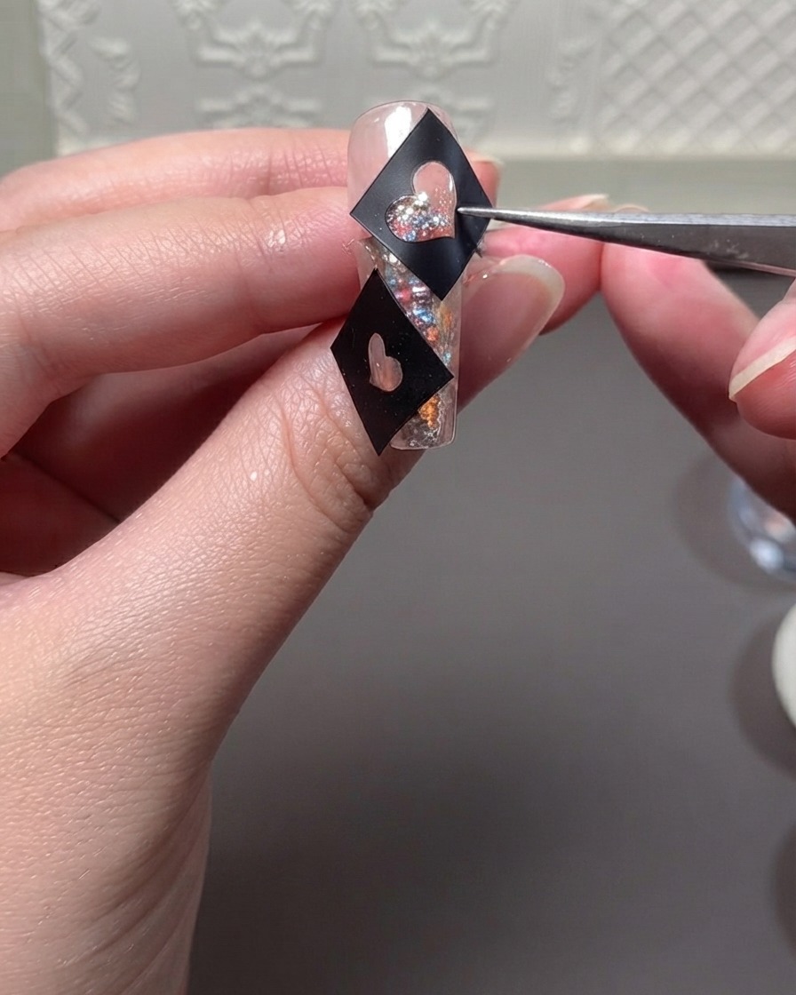 Unas pinzas sosteniendo una plantilla de corazón negro sobre una uña con glitter mientras se aplica gel.