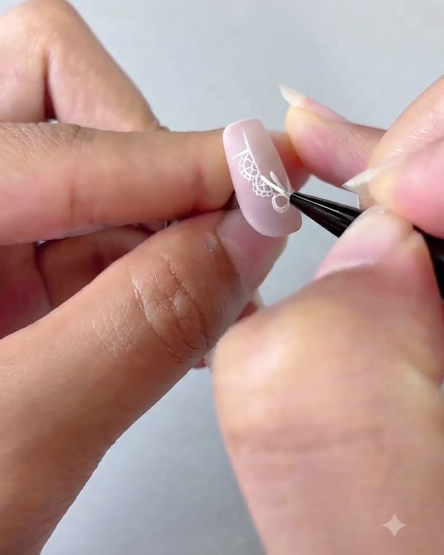 Un pincel para nail art añadiendo puntos blancos detallados y una estrella de cuatro puntas sobre una uña con degradado rosa.