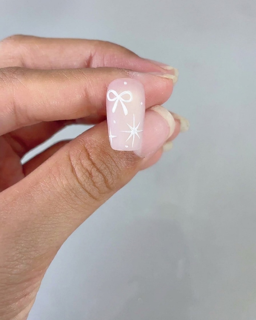 Un diseño de uña terminado que presenta un lazo blanco y una estrella de cuatro puntas sobre una base degradada rosa sostenida para su visualización.