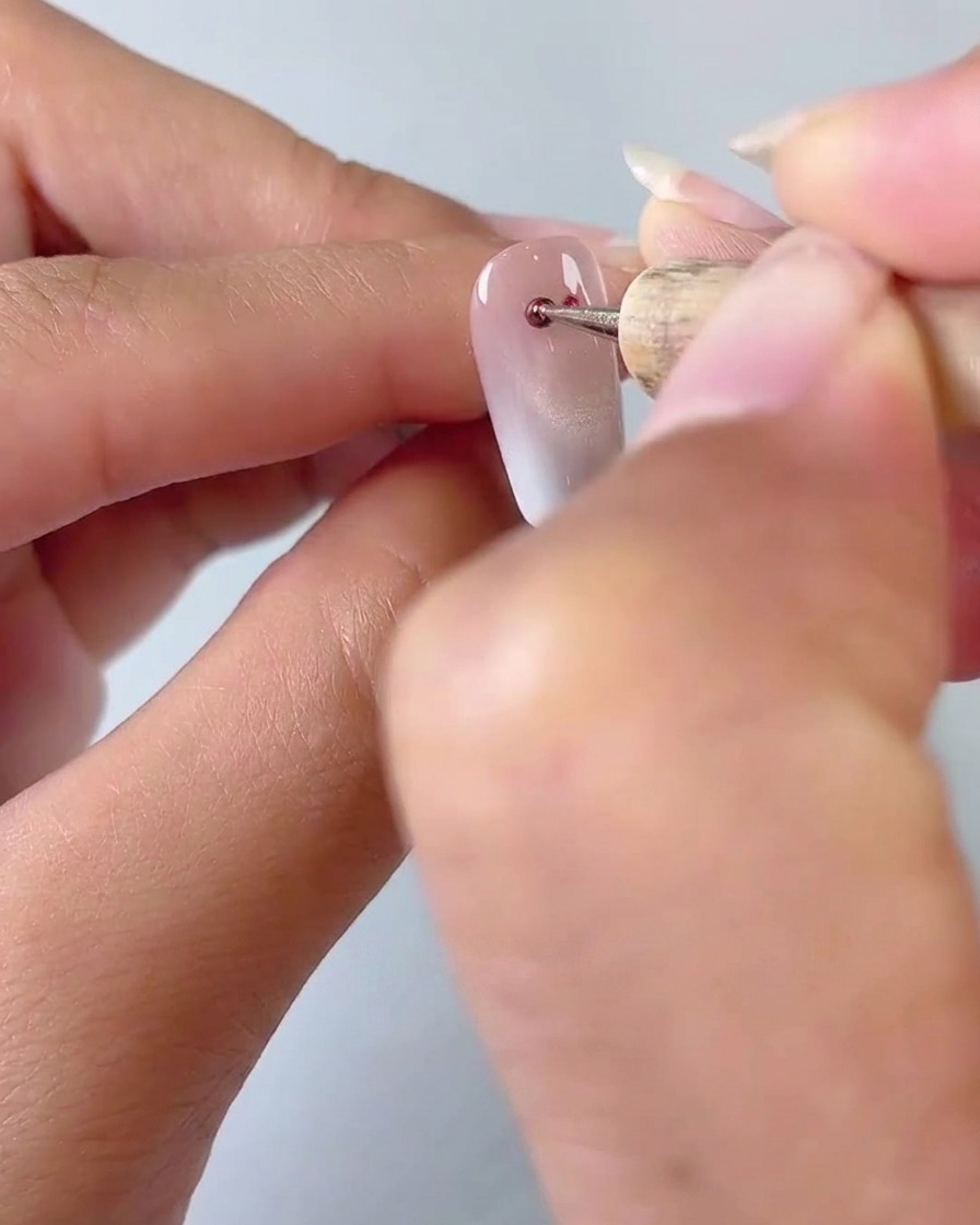Un primer plano de una herramienta de punteado aplicando dos pequeños puntos de gel ojo de gato rojo sobre una base de uña con degradado lechoso.
