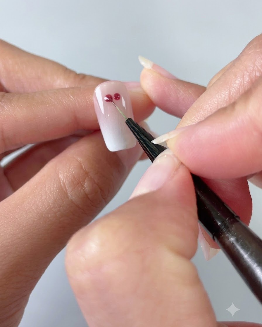 Un pincel fino de nail art conectando dos puntos de gel rojo en forma de corazón sobre una uña degradada con ojo de gato.