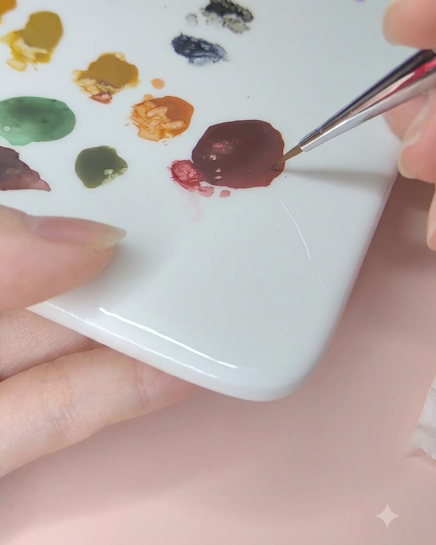 Un pincel fino para nail art recogiendo pintura de acuarela rojo oscuro de una paleta de cerámica.