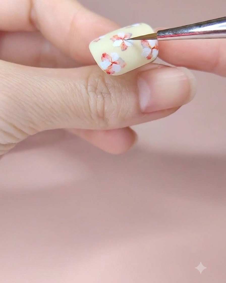 Un pincel fino de nail art añadiendo un toque de acuarela roja al centro de una flor blanca en una uña amarillo pálido.