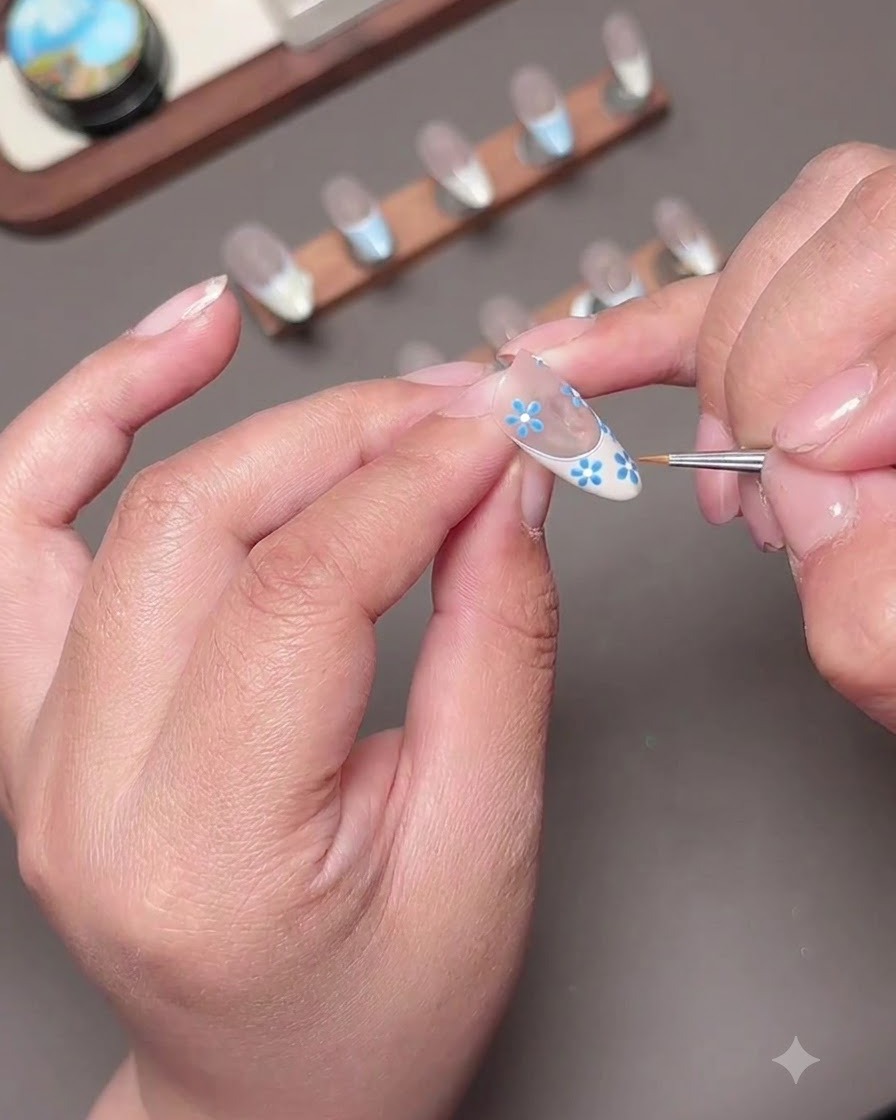 Una toma macro de pequeños pétalos azules siendo pintados en la superficie de la uña con un pincel de detalle.
