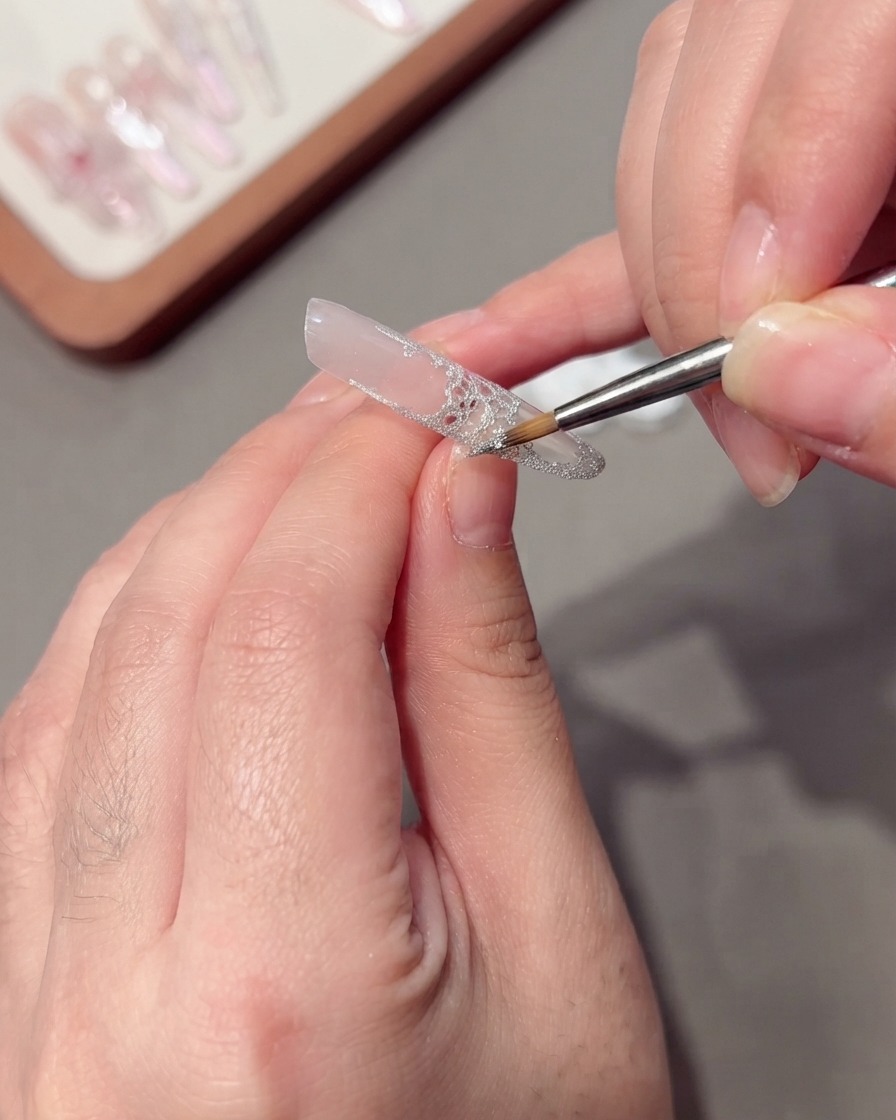 Un pincel fino aplicando spider gel plateado brillante a lo largo del borde de un diseño de encaje.