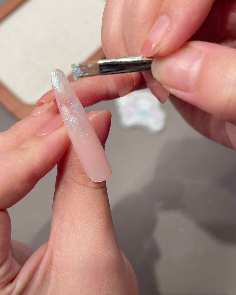Un pincel aplicando una capa húmeda de base coat sobre la superficie de una uña larga y rosa.