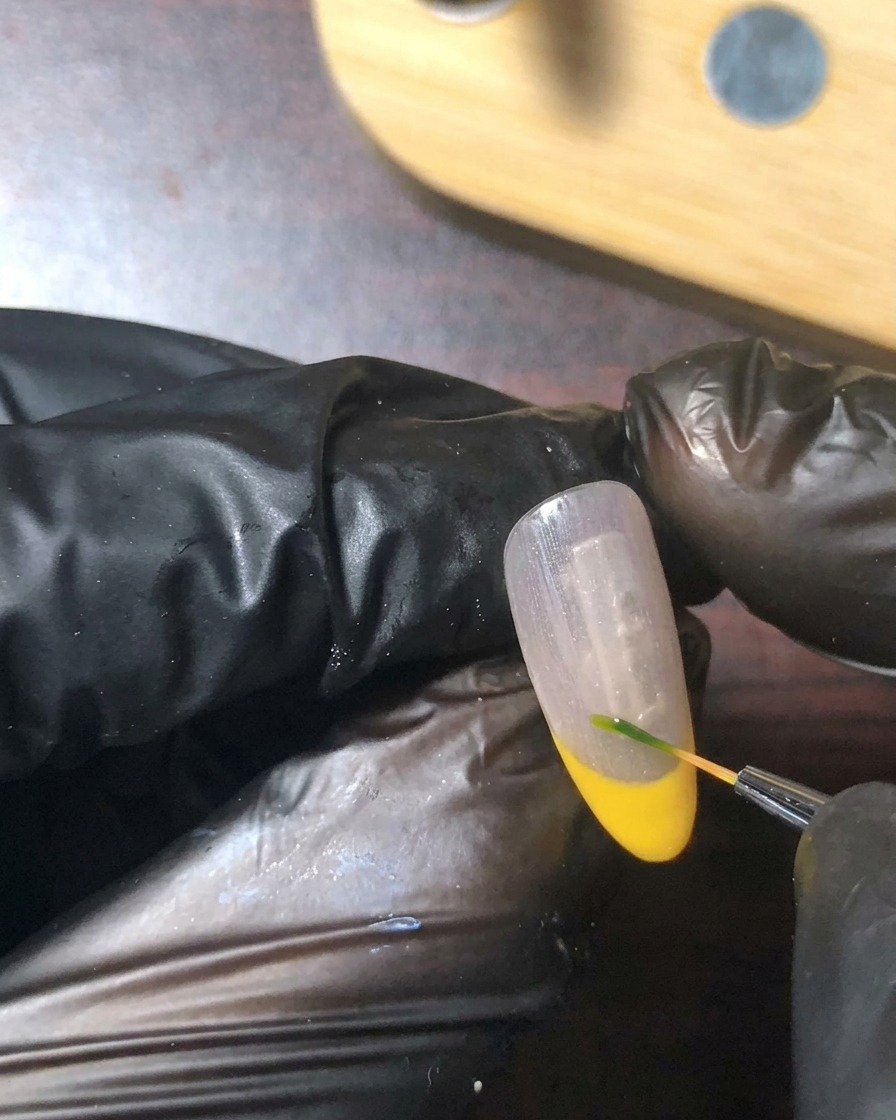 Un pincel de detalle fino pintando un tallo y una hoja verde diminutos cerca de la línea de la sonrisa de la uña.