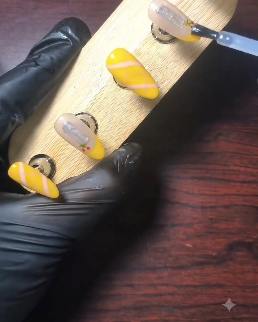 Una mano enguantada utilizando un pincel para aplicar una capa superior transparente y brillante sobre una uña decorada con punta francesa amarilla en un soporte de exhibición de madera.