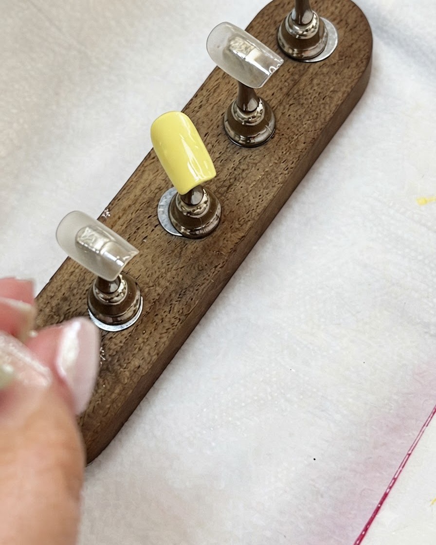 Tips de uñas en un soporte de madera mostrando una uña de color amarillo lechoso y otras en tono nude.