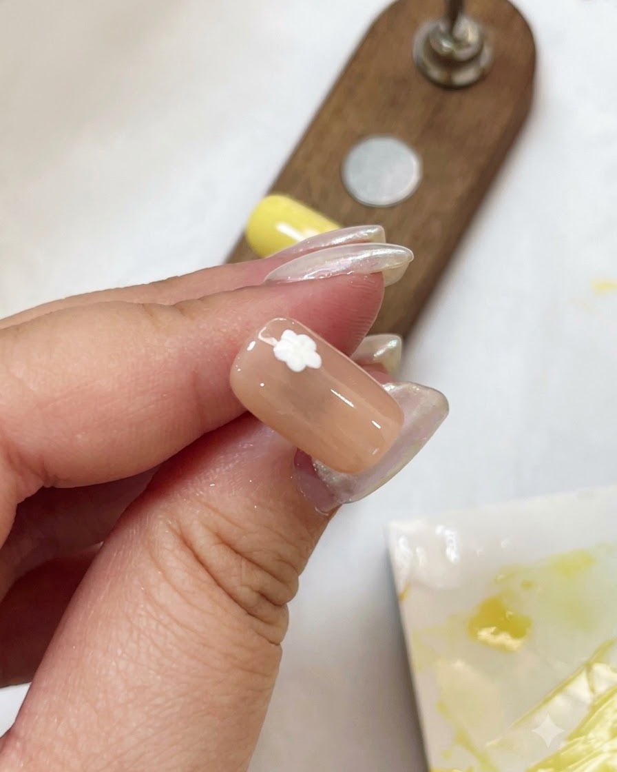 Un primer plano de una uña nude con un diseño de flor blanca curándose con una pequeña lámpara UV portátil violeta.