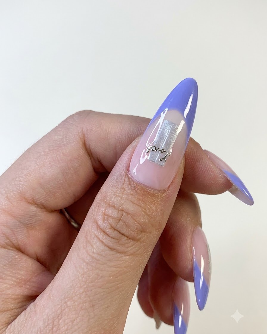 Una vista de cerca de una uña de pulgar que presenta una punta francesa azul, un elemento base de papel de aluminio plateado y una delicada pegatina de letras.