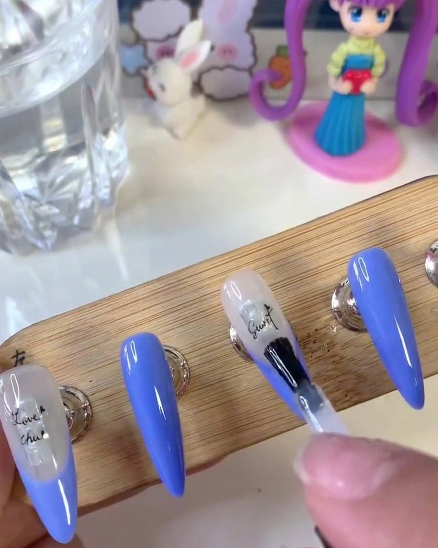 Un pincel de uñas aplicando una capa transparente de gel de refuerzo sobre la superficie de una uña con pegatinas de letras.