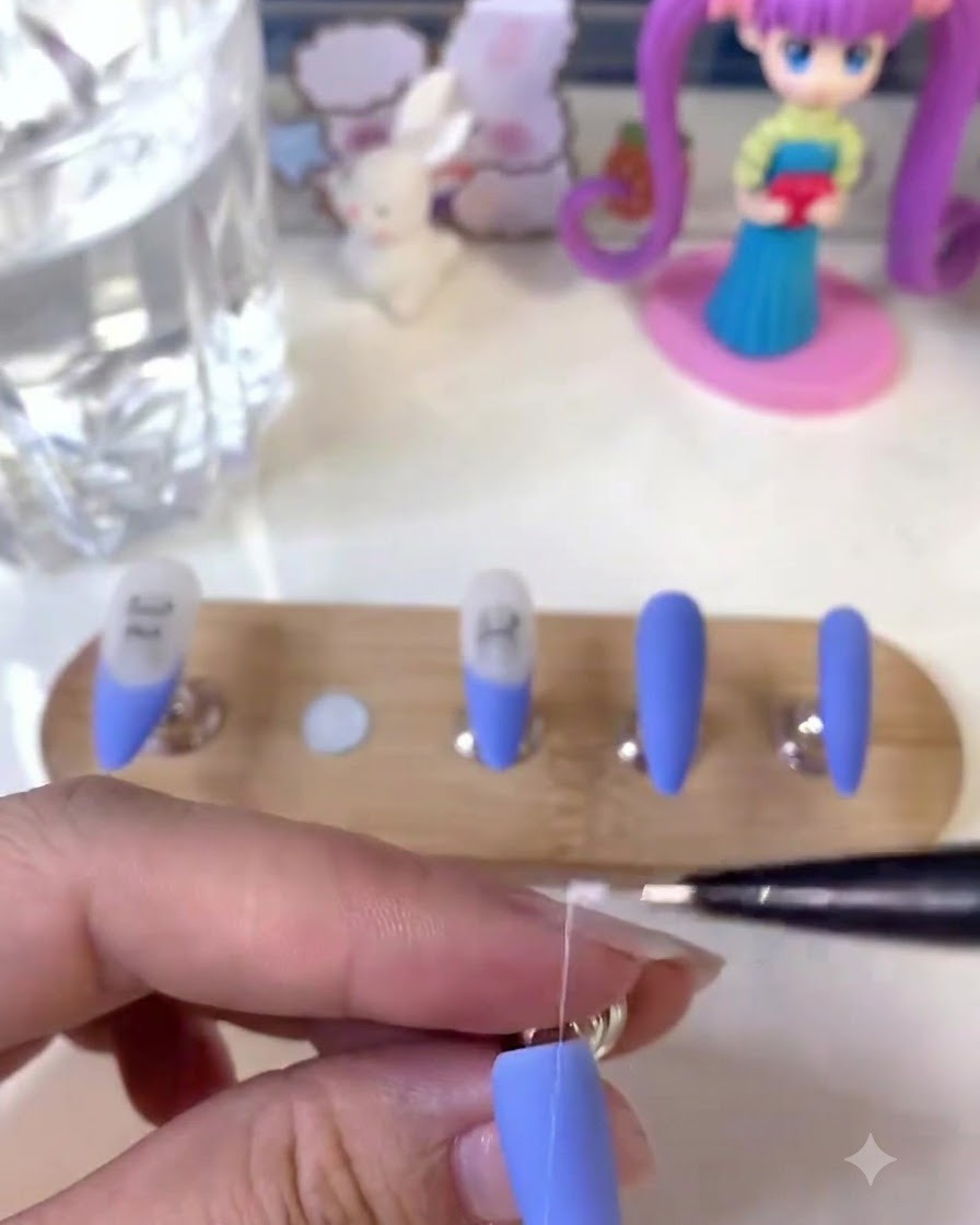 Una artista de uñas sosteniendo un pincel delineador fino y dibujando líneas blancas detalladas en una uña azul mate.
