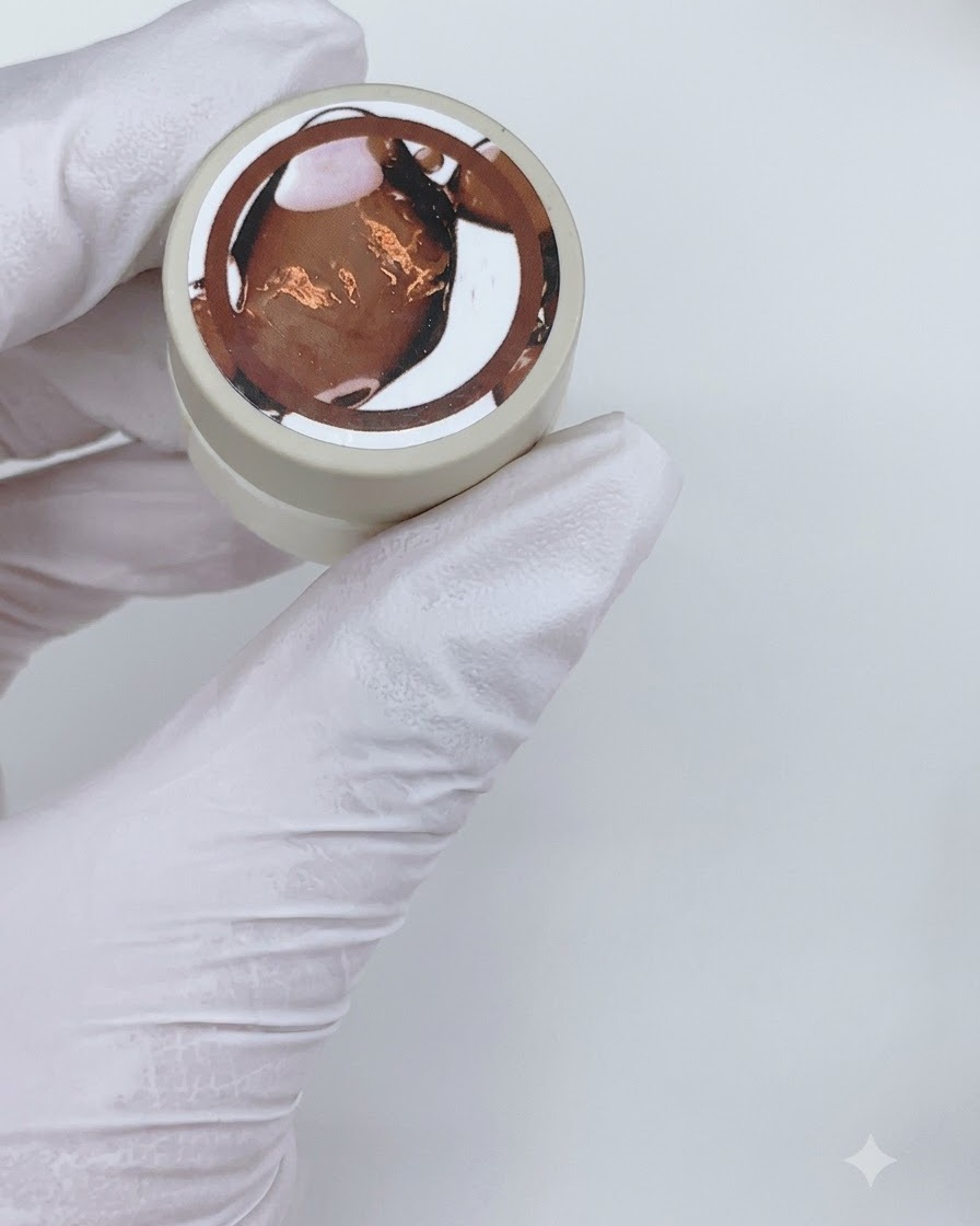 Un pincel de gel plano aplicando una capa de esmalte en gel marrón translúcido sobre un bastón de exhibición de uñas transparente.