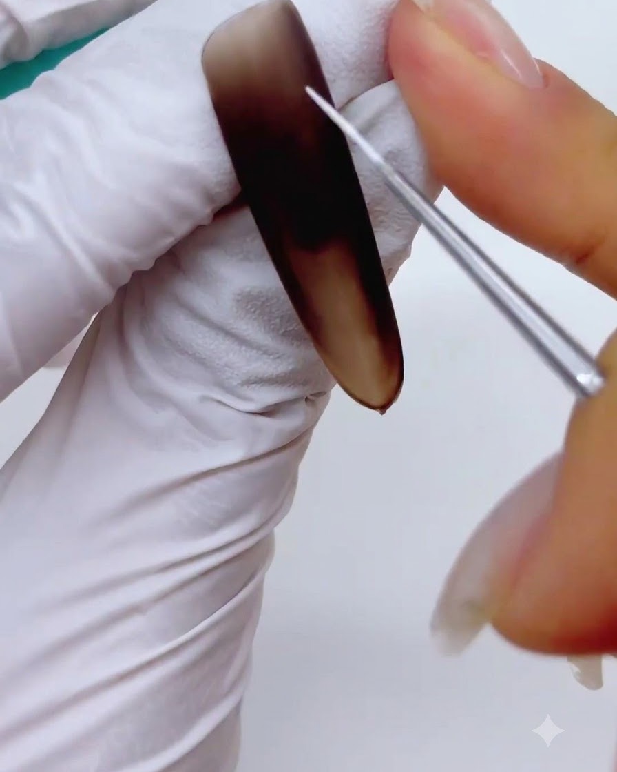 Un primer plano de un pincel delineador fino para arte en uñas pintando líneas blancas finas y entrecruzadas para formar un esqueleto de copo de nieve sobre una uña marrón mate.