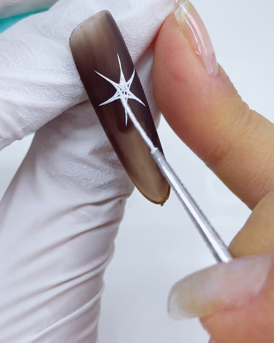 Un primer plano de un pincel de detalle fino conectando las puntas y rellenando las secciones centrales de un diseño de estrella blanca sobre una base de gel marrón mate.