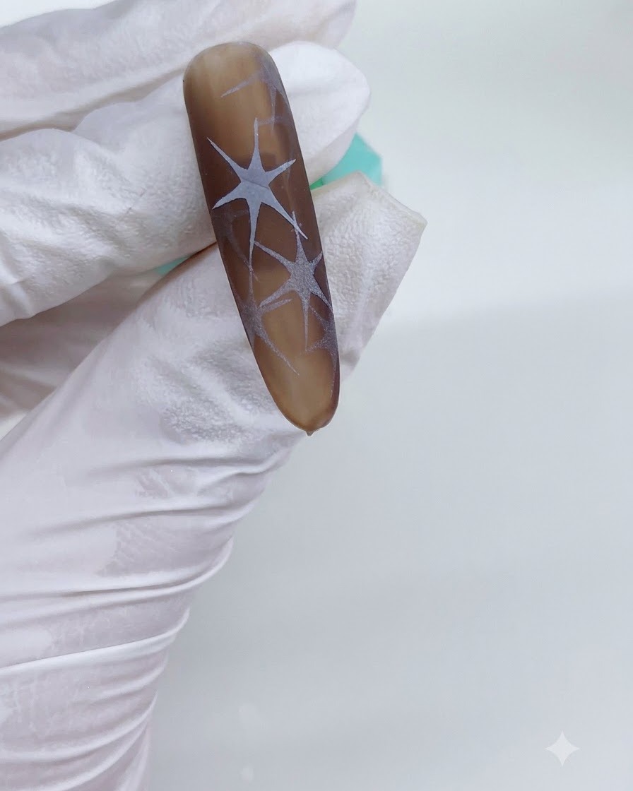 Una mano con guantes sosteniendo una uña con tonos marrones bajo una lámpara UV con patrones de estrellas blancas curadas visibles.