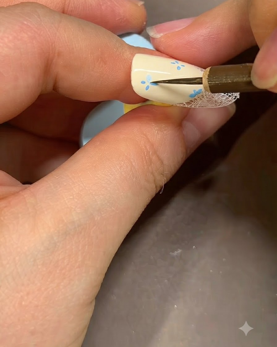 Un pincel fino de nail art agregando acentos azules detallados al centro de una flor pintada en una uña beige.