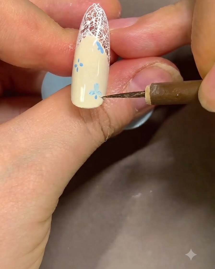 Primer plano de una manicurista usando un pincel fino para limpiar los bordes de un diseño de flor azul cerca de la punta francesa.