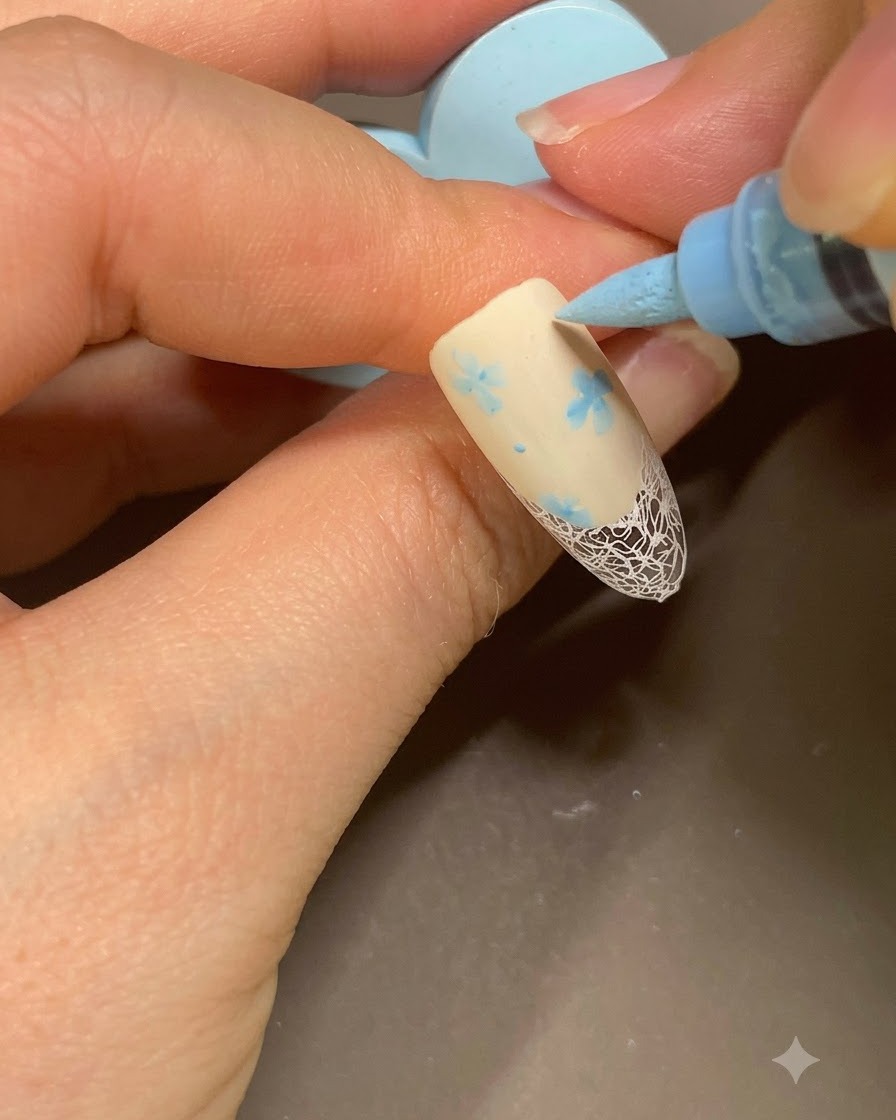 Un marcador azul de nail art agregando puntos finales junto a un patrón floral en una manicura francesa beige.