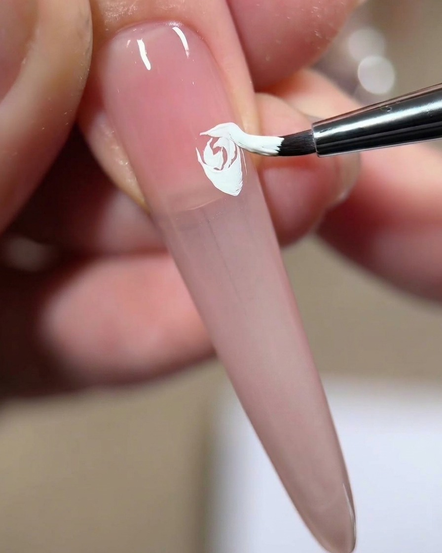 Un pincel de uñas presionando y arrastrando gel blanco para añadir un cuarto pétalo en capas a una rosa blanca pintada sobre una uña rosa.