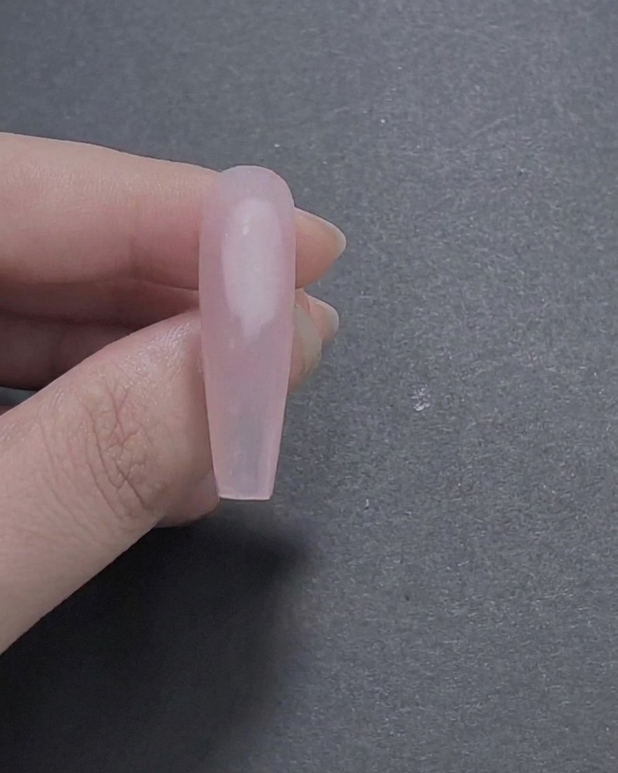 Un primer plano de una uña larga en forma de ataúd pintada con una capa base de gel rosa translúcido suave y brillante, sostenida entre los dedos.