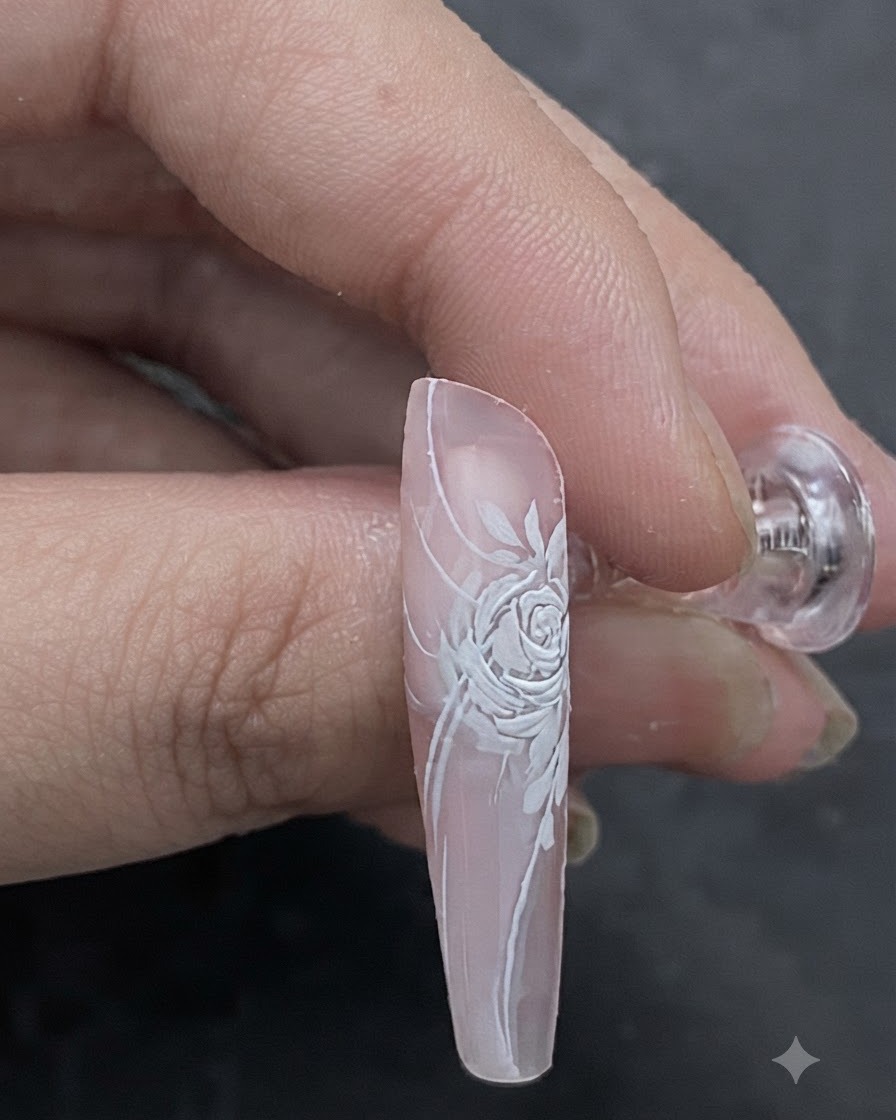 Una imagen de cerca que muestra un tono rosa claro siendo aplicado con un pincel en el centro de un diseño dimensional de rosa blanca en las uñas.