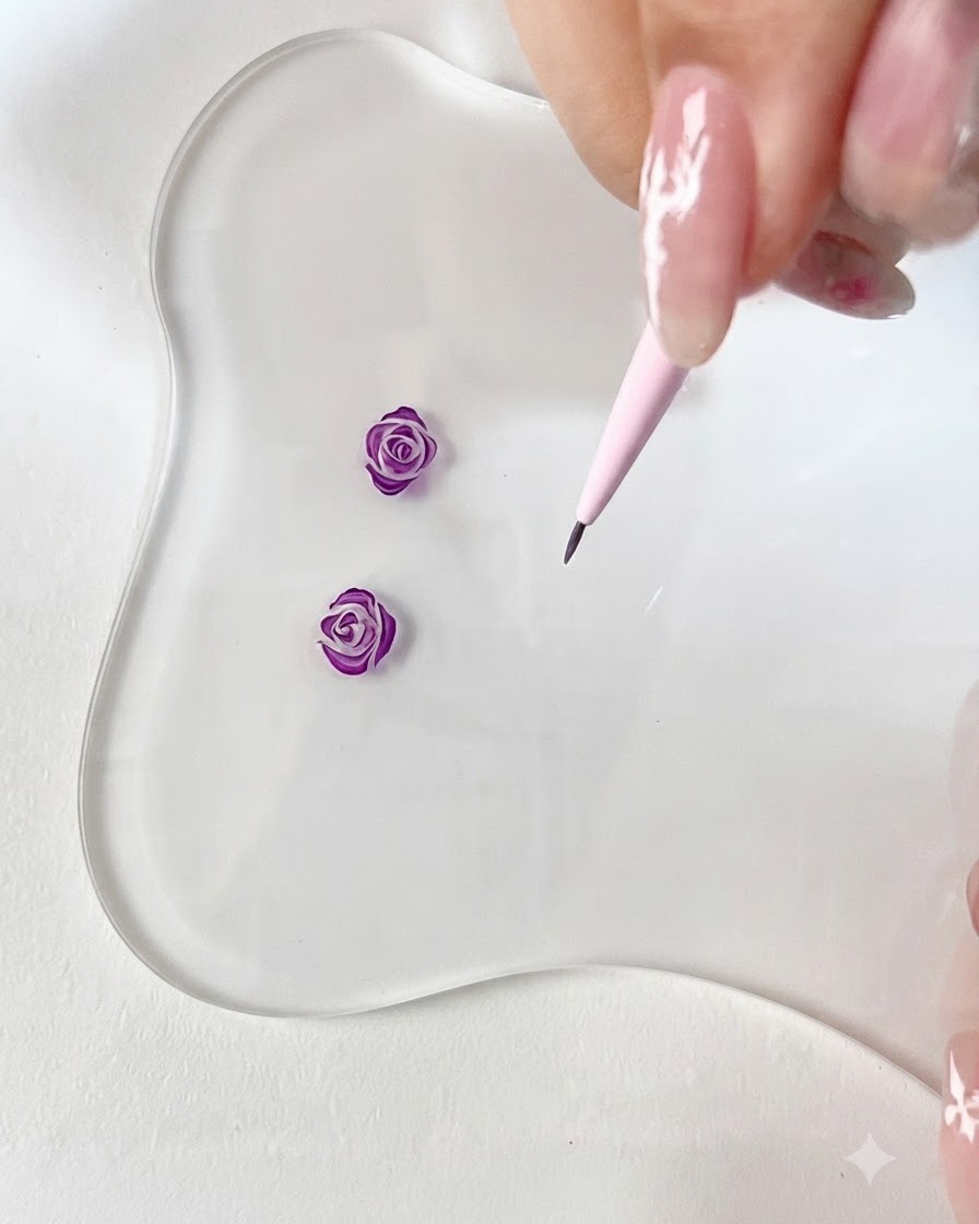 Vista cercana de un pincel delineador fino dibujando un pequeño gancho de pétalo morado curvo sobre una paleta transparente.