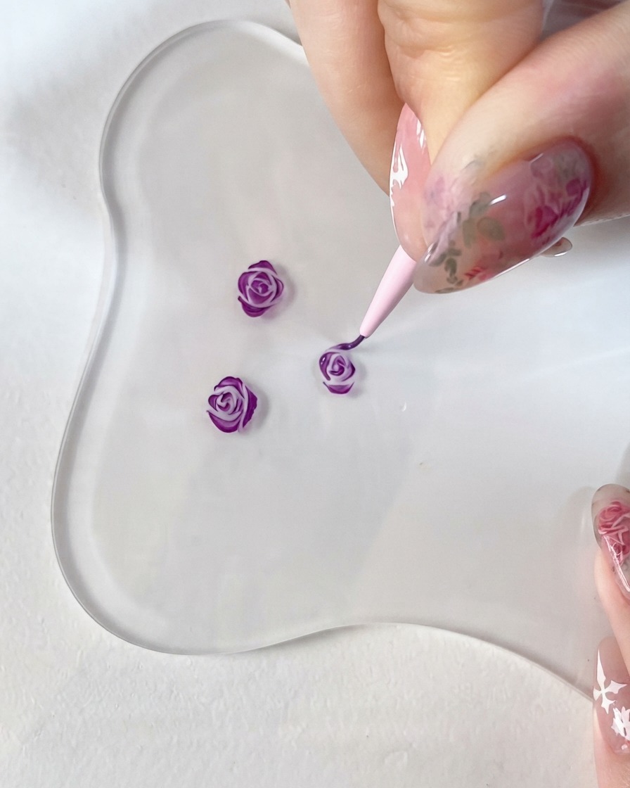 Un primer plano de una mano sosteniendo un pincel de detalle fino, pintando meticulosamente acentos de color morado oscuro en un pequeño diseño de rosa en 3D sobre una paleta de vidrio transparente.