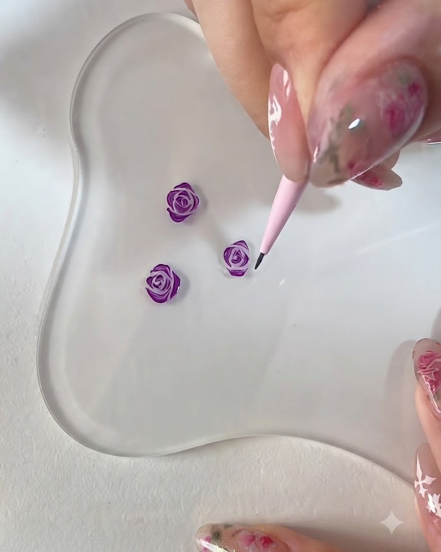 Un primer plano de una mano usando un pincel de detalle rosa para refinar los bordes de una rosa morada pintada en 3D sobre una paleta transparente.