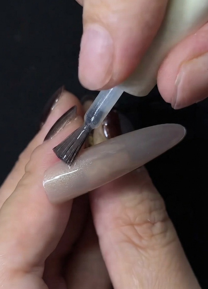 Un pincel de uñas aplicando esmalte de gel cat eye transparente sobre la superficie de una extensión de uña nude.