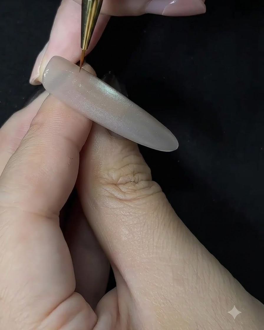 Un primer plano de una artista de uñas usando un pincel de detalle fino para aplicar gel blanco con un movimiento de punteado sobre una extensión de uña esculpida.