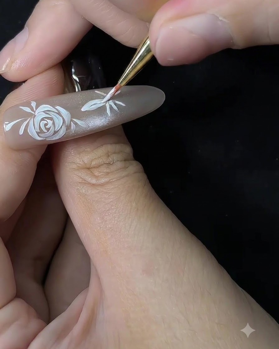 Una toma de cerca de un pincel fino comenzando a dar forma a los pétalos de una flor secundaria en una extensión de uña de color nude.