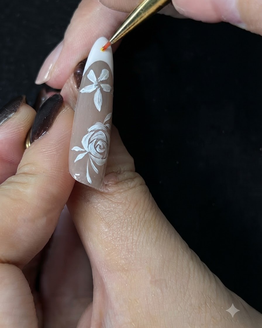 Un primer plano de una mano usando un pincel delineador de detalle fino para pintar pequeñas enredaderas de acento blancas alrededor de una rosa blanca pintada a mano en una uña de gel nude.