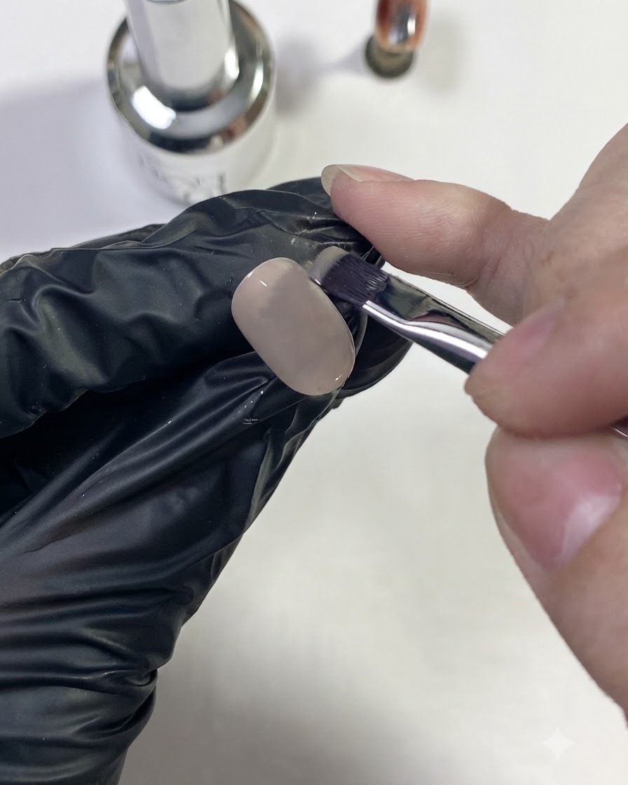 Una mano con guantes aplicando un color base nude sobre la punta de una uña.