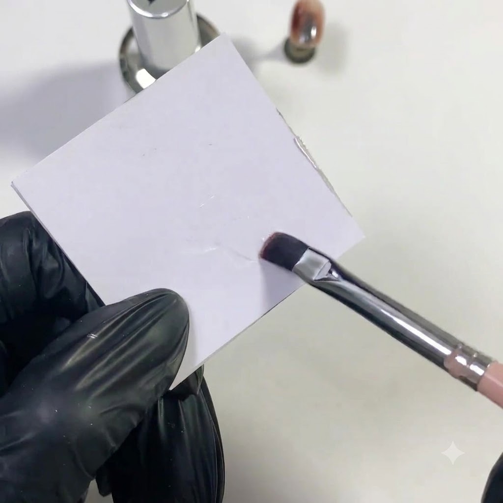 Un pincel de uñas plano aplicando base en gel transparente sobre la superficie de una paleta blanca.