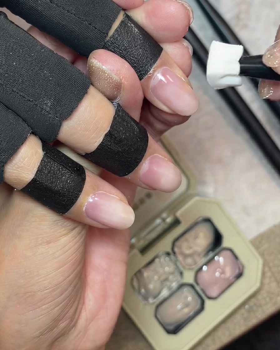 Una herramienta de esponja aplicando gel cover nude lechoso a lo largo del borde libre natural de una uña, con una paleta de gel al fondo.