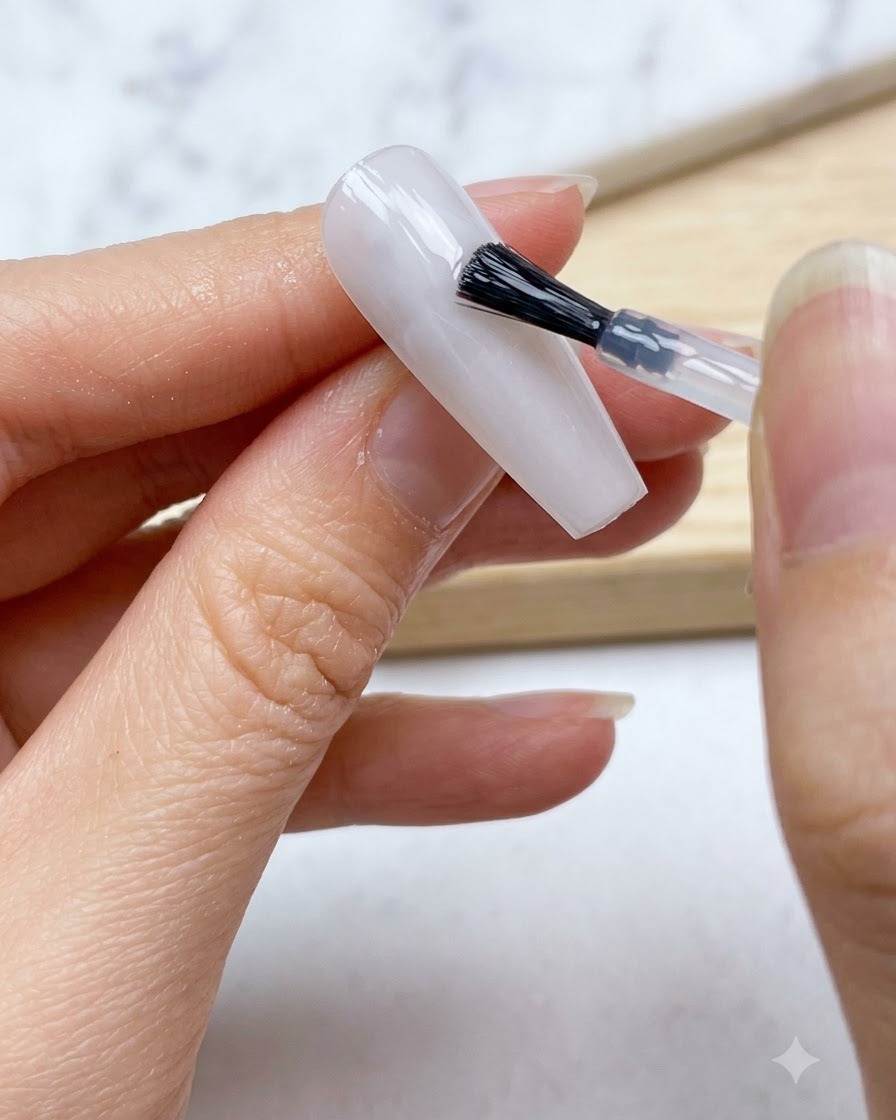 Un pincel de uñas aplicando una capa suave de base de gel blanco lechoso translúcido sobre toda la uña.