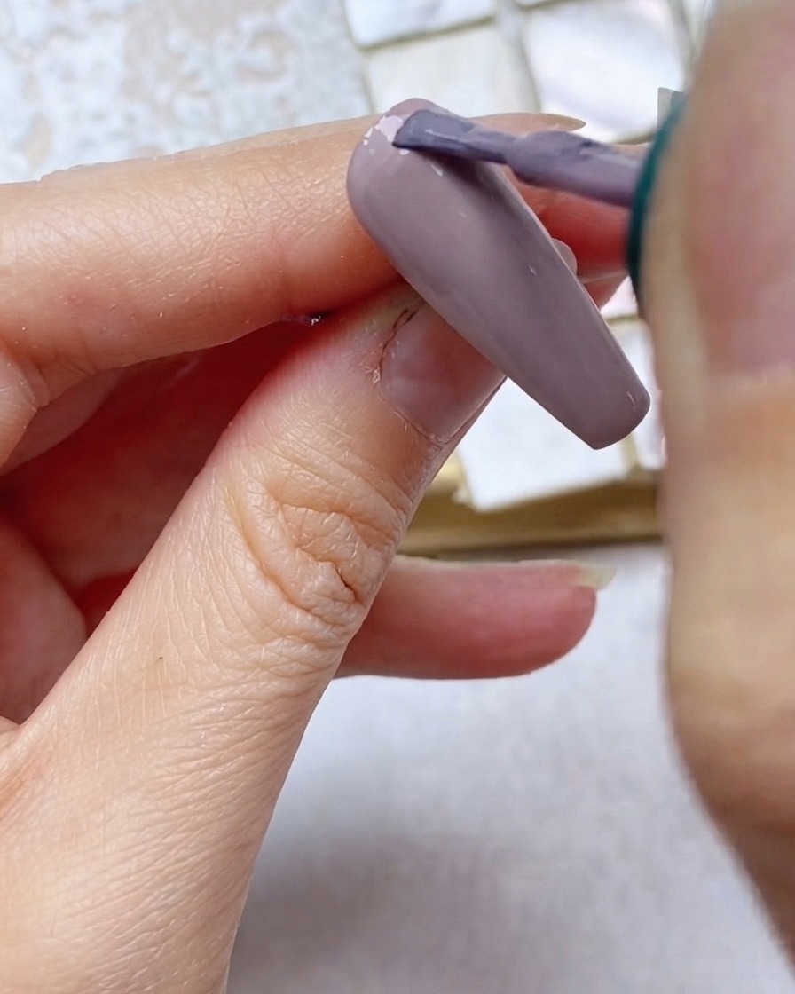 Un pincel aplicando esmalte en gel gris morado en el tercio superior de la uña cerca de la cutícula.