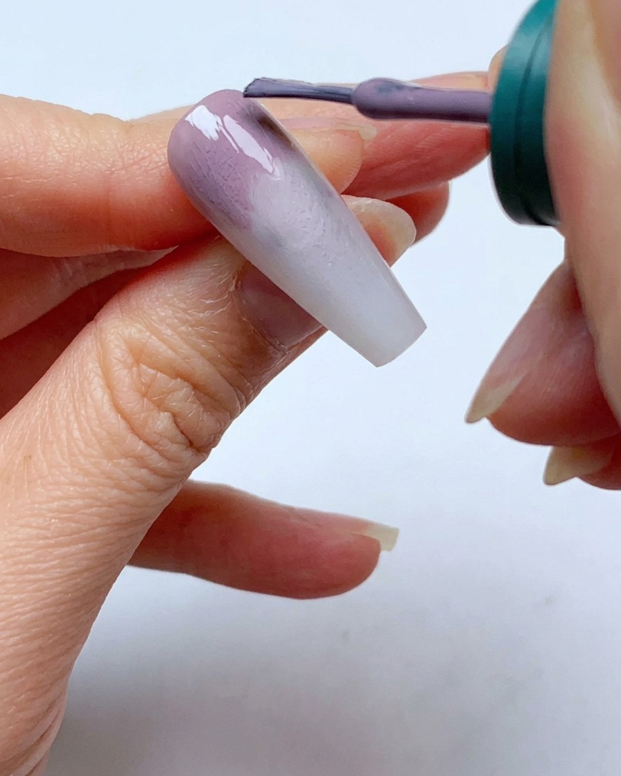 Un pincel de esmalte de uñas aplicando una segunda capa de gel gris morado cerca de la cutícula de una uña.