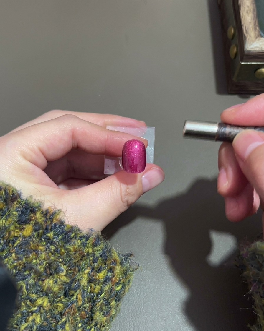 Una mano sosteniendo un imán junto a una uña cat-eye morado oscuro para formar un brillo.