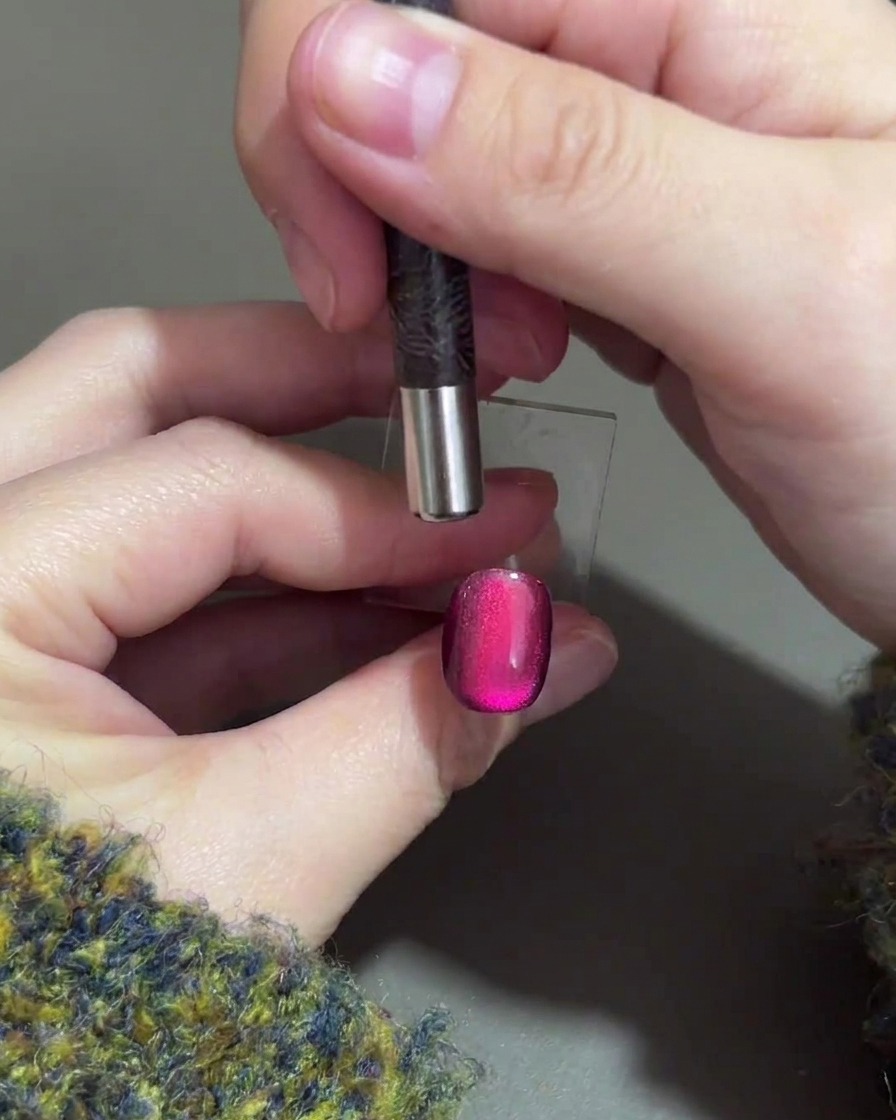 Una mano sosteniendo un imán cilíndrico sobre el borde superior de una uña de gel morada para empujar el brillo magnético hacia abajo.