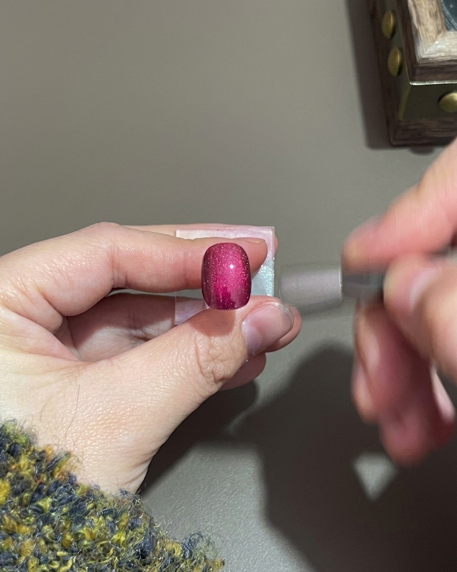 Una mano sosteniendo un pequeño imán cilíndrico cerca de una uña de gel cat-eye morada.