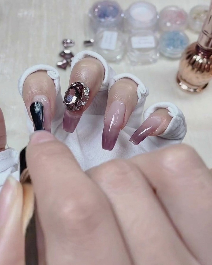 Una artista de uñas aplicando el top coat alrededor de la base de una gema de cristal en una uña con degradado terminada.